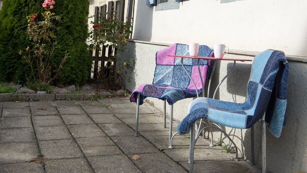 Two metal chairs with colorful blankets, a stone pathway, and a building with blue shutters.