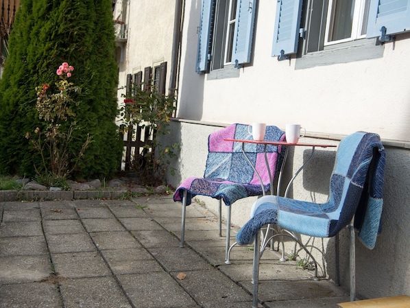 Two metal chairs with colorful blankets, a stone pathway, and a building with blue shutters.
