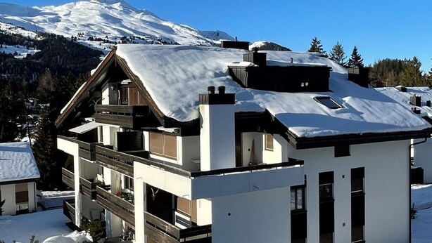 Een moderne, tweelaagse chalet met een met sneeuw bedekte dak, tegen een achtergrond van een besneeuwde berg.