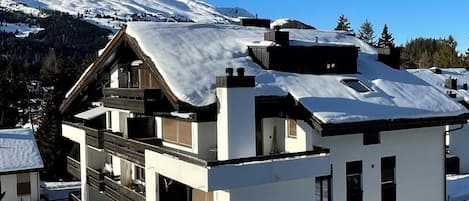 A modern two-story chalet with a snow-covered roof, set against a backdrop of a snow-capped mountain.