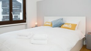 Een net opgemaakt bed met witte lakens en twee kussens met de tekst 'smile', een bedlamp en een raam met uitzicht op gebouwen.