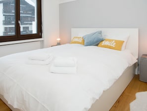 A neatly made bed with white linens and two pillows with 'smile' text, a bedside lamp, and a window with a view of buildings. - 1619 Sundroina Prada C by Interhome (Lenzerheide)