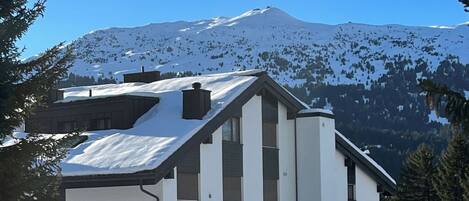 A modern building with a sloped roof, large windows, and a snow-covered landscape with mountains in the background.
