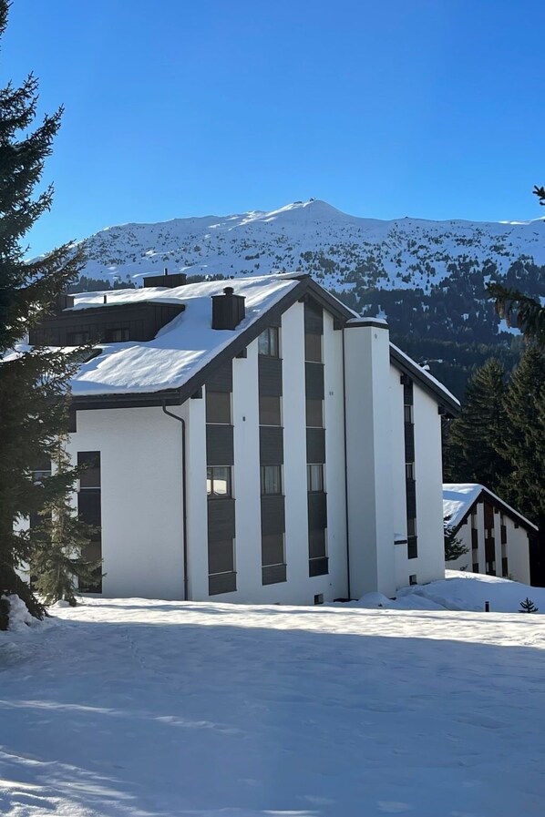 A modern building with a sloped roof, large windows, and a snow-covered landscape with mountains in the background.