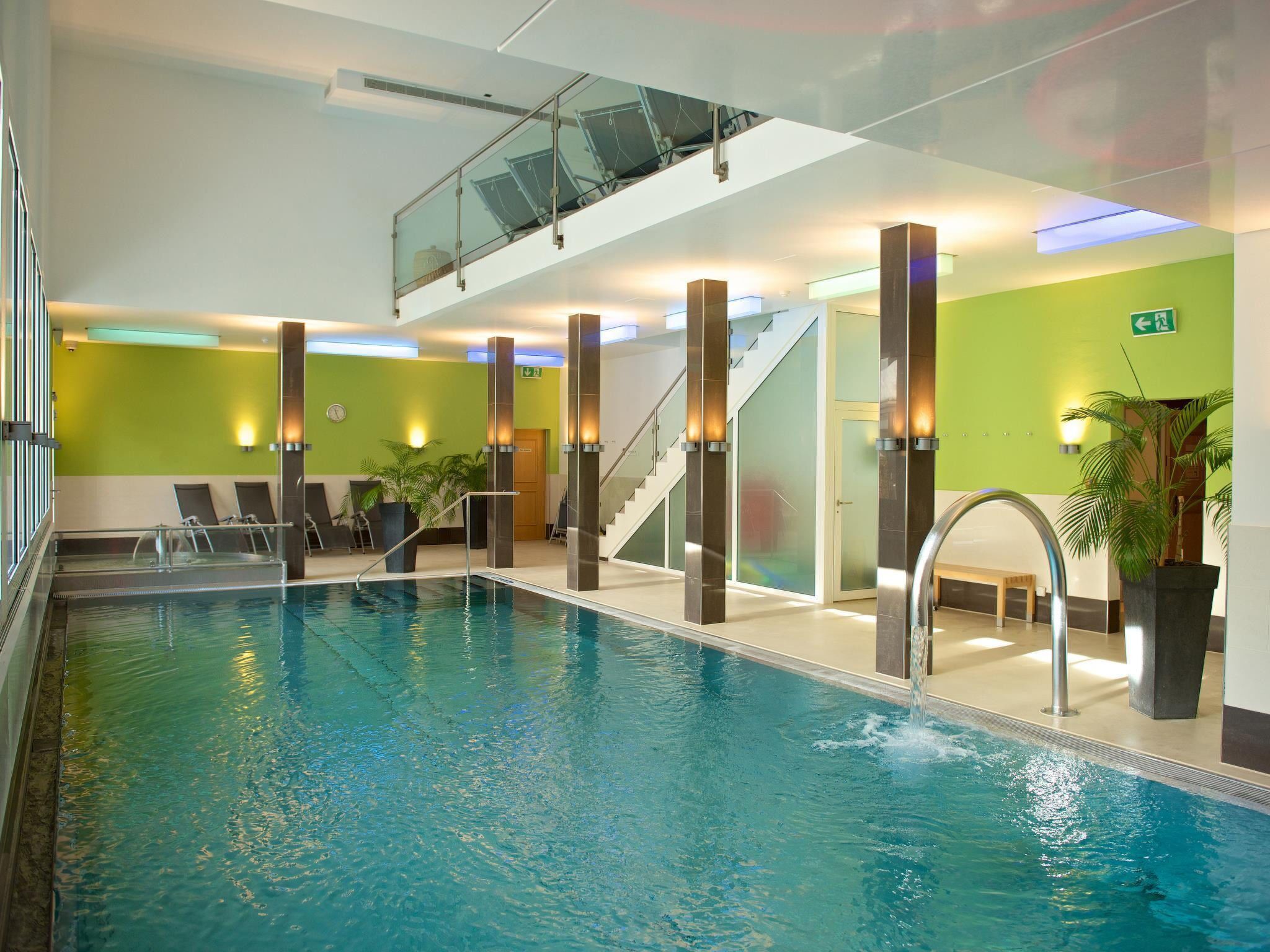 An indoor swimming pool with clear water, surrounded by walls with green panels and modern lighting fixtures.