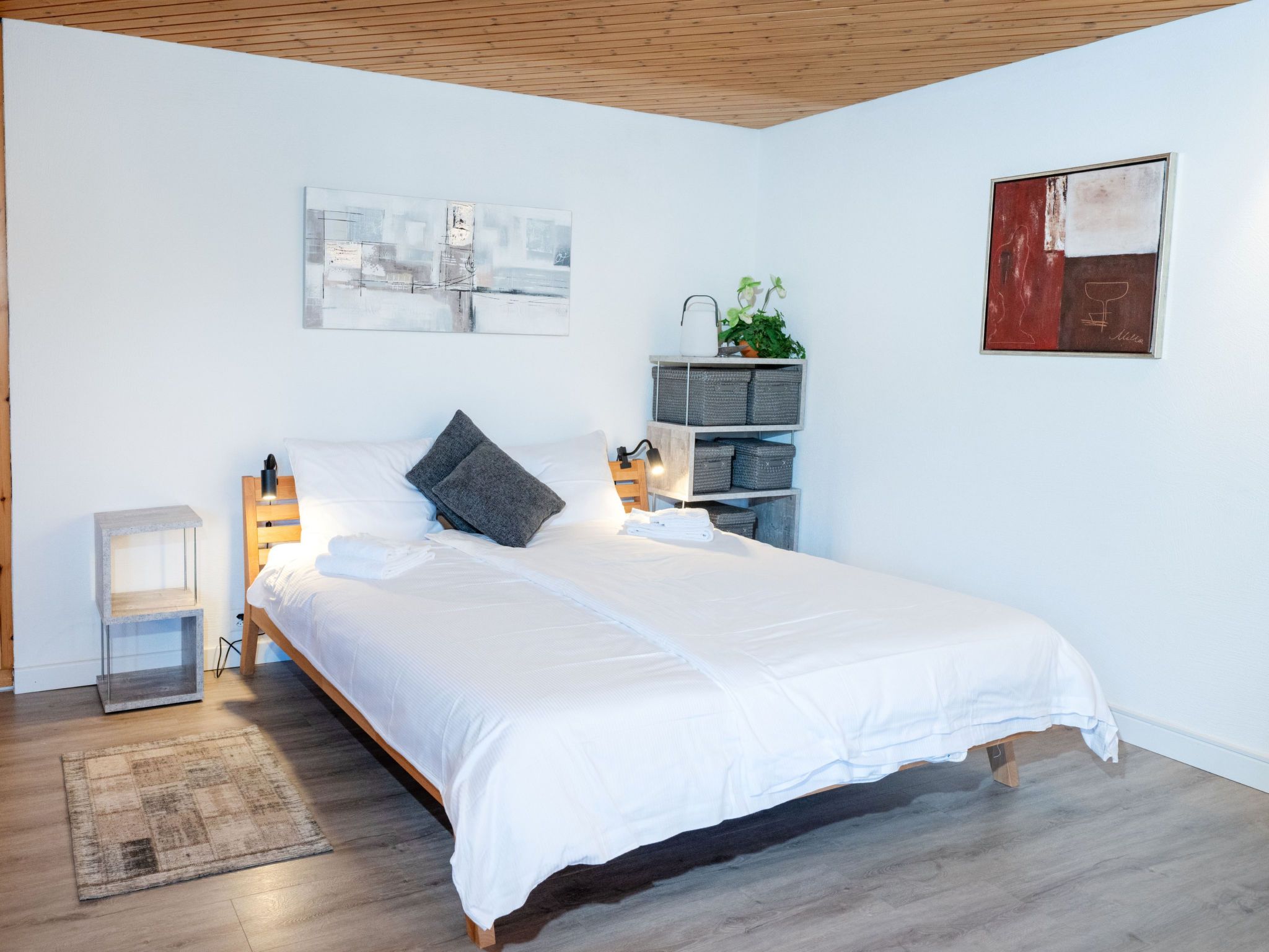Ein modernes Schlafzimmer mit einem hölzernen Bett, weißer Bettwäsche, einem Nachttisch mit Lampe, einer Wandablage mit Pflanze und Kunstwerken.