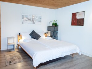 Una camera da letto moderna con un letto di legno, biancheria bianca, un comodino con una lampada, uno scaffale a parete con una pianta e opere d'arte.