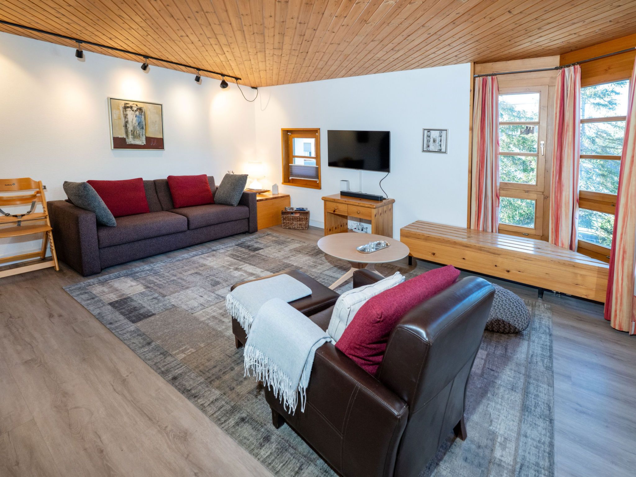 A modern living room with a sofa, a leather couch, a coffee table, and a TV. There are windows with curtains and a wooden ceiling.