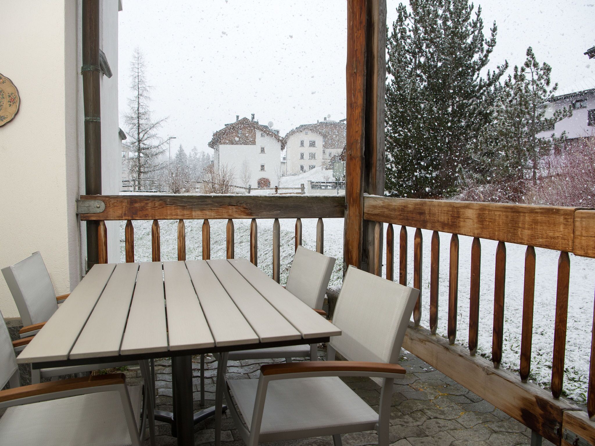Una terraza de madera con una mesa y sillas, con vista a un paisaje nevado y edificios.