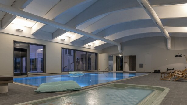 An indoor swimming pool with a tiled floor, a lounge area with chairs, and a large window. - 1609 Schweizerhof 604 by Interhome (Lenzerheide)