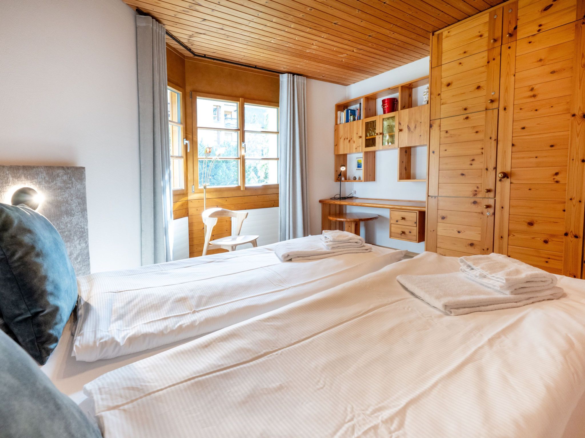 A hotel room with two beds, a wooden wardrobe, a desk, and a window with curtains.