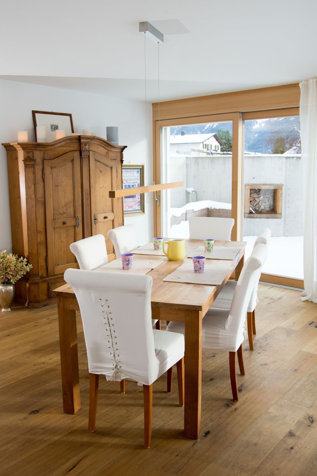 Un coin repas avec une table en bois et des chaises blanches, un buffet en bois et une fenêtre donnant sur un paysage enneigé.