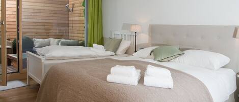 Cama doble con colcha y almohadas color beige, mesita de noche con lámpara y vista a una cabaña de madera a través de un vidrio.