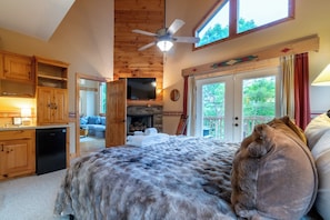 Ein Schlafzimmer mit einem großen Bett, einem Deckenventilator, einem Fernseher, Holzelementen, einem Kamin und einem Balkon mit Vorhängen.