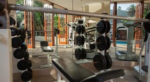 A gym with weightlifting equipment, a bench, and a view of a pool and greenery. - The Wilderness Hotel (Wilderness)