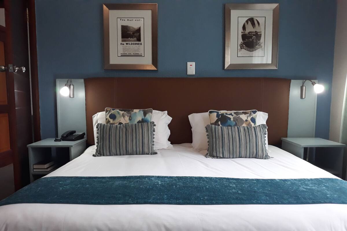 A hotel room with a large bed, two framed pictures on the wall, a nightstand with a phone, and a bedside table with a lamp.