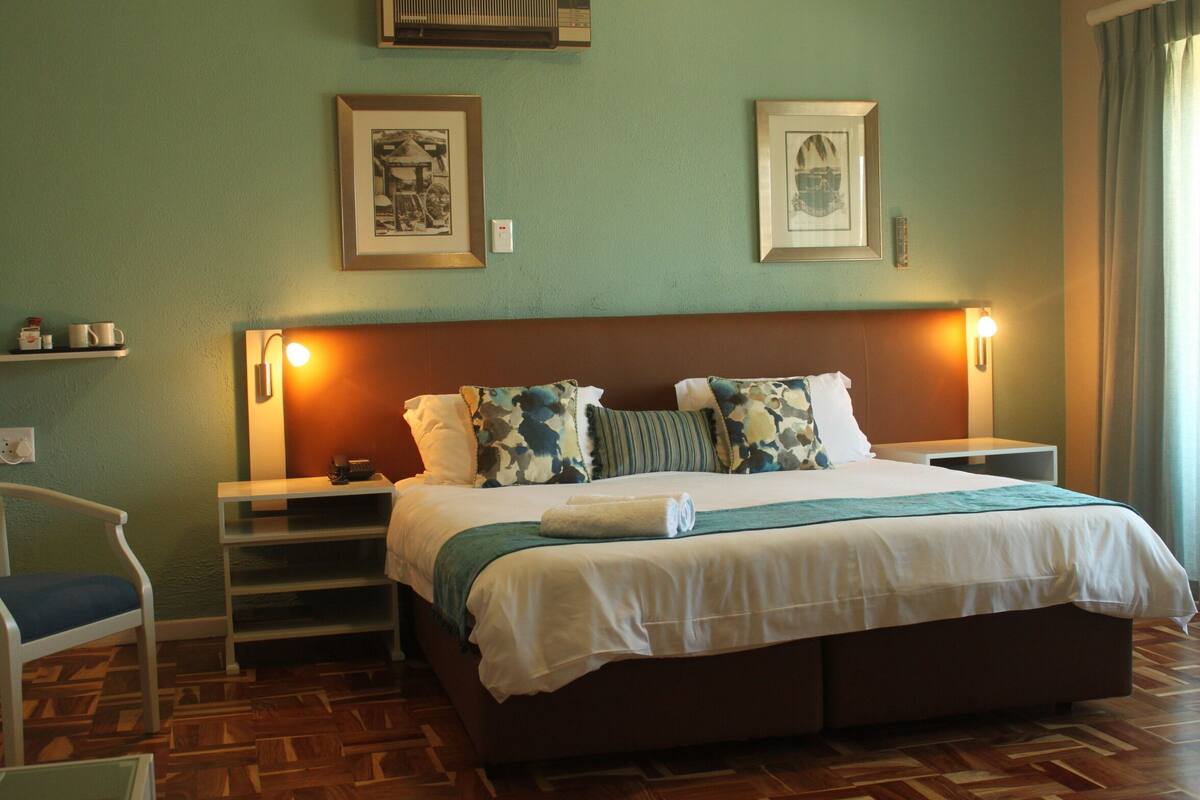 A neatly arranged hotel room with a large bed, bedside tables, a chair, and a shelf with cups.