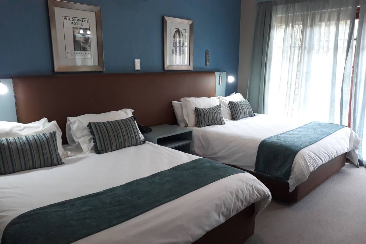 A hotel room with two beds, each with white bedding and striped pillows, a brown headboard, and a framed picture of the Wilderness Hotel on the wall.