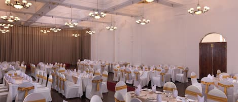 A banquet hall with round tables set for dining, white chairs with gold sashes, and chandeliers hanging from the ceiling.