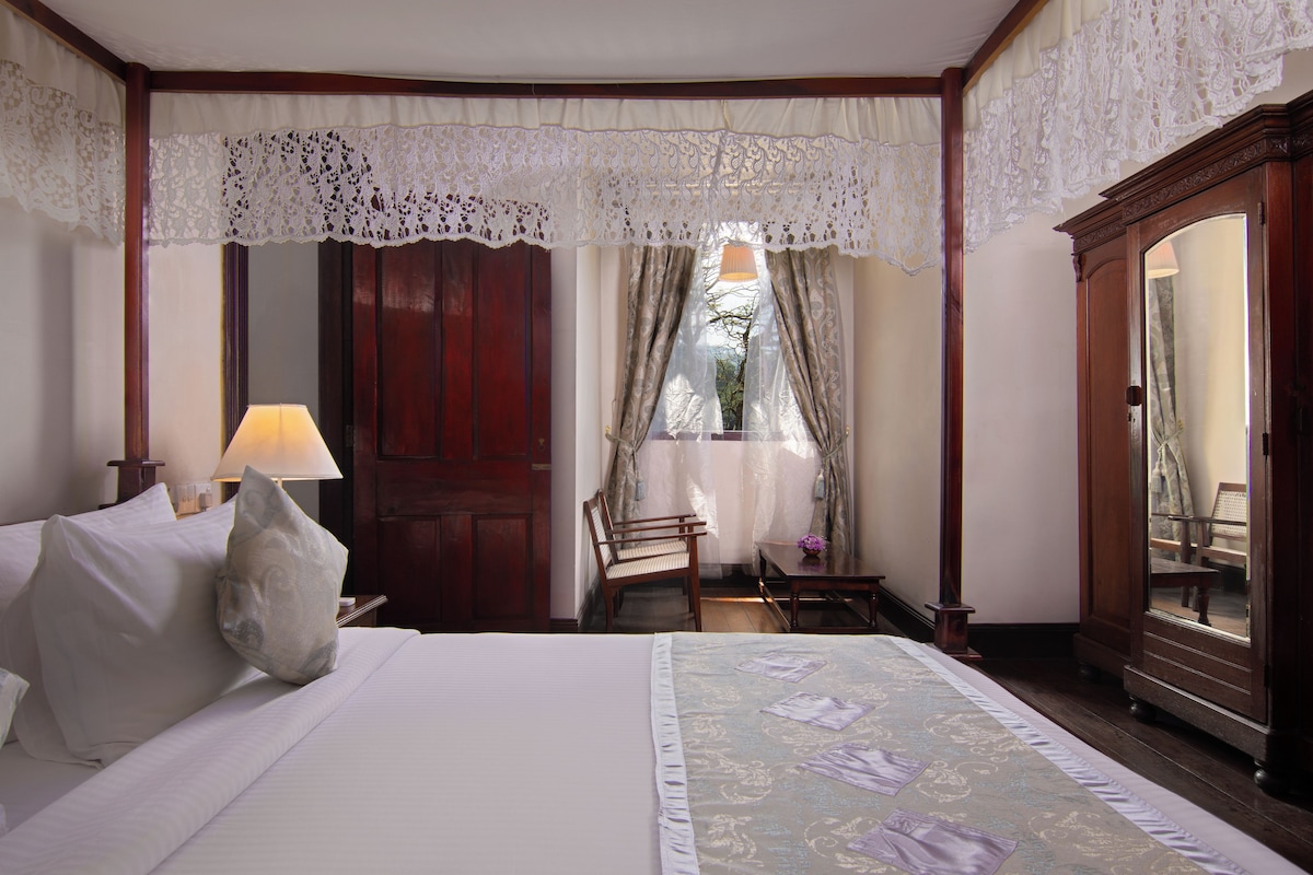 Un cuarto de hotel ordenado con una cama grande, una mesita de noche de madera, un escritorio con una lámpara y una silla.
