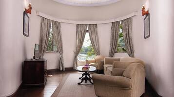 Una habitación con techo curvo, una mesa redonda con un libro y un florero, dos sillones color beige, un aparador de madera y vistas a la vegetación a través de amplias ventanas con cortinas.