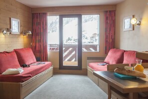 Una habitación con pared de madera, una ventana con vistas a un paisaje nevado, un sofá con cojines rojos, una mesa con un vaso de zumo de naranja y un par de auriculares sobre el sofá.