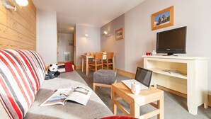 A cozy living room with a striped sofa, a small table with a laptop, a TV on a stand, and a dining area in the background. - Residence Pierre et Vacances Balcons de Bellevarde (Val-d'Isere)