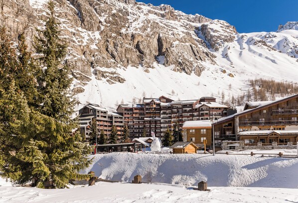Un centro turístico en la montaña cubierto de nieve, con edificios de madera, un telesilla y un gran pino.