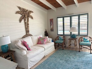 Ein weißes Sofa mit bunten Kissen, ein blaues gemustertes Teppich, Korbstühle mit türkisen Kissen, eine Holzbalkendecke und ein Fenster mit Jalousien.