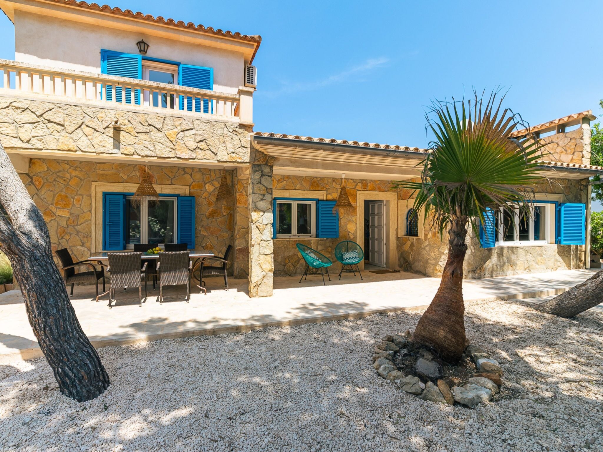 A two-story building with stone walls and blue shutters, outdoor seating with chairs, and a palm tree.