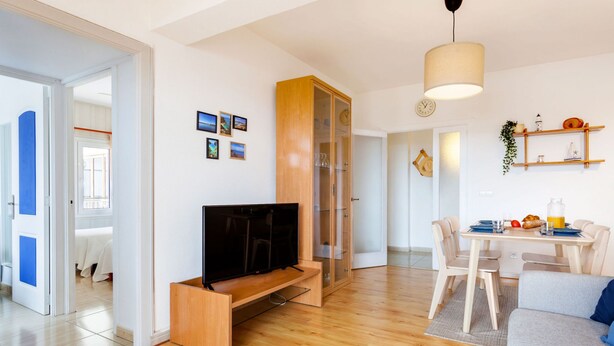 Un salon moderne avec un téléviseur à écran plat posé sur un meuble en bois, un coin repas avec une table et des chaises, et un espace cuisine avec un luminaire suspendu.