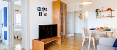 A modern living room with a flat-screen TV on a wooden stand, a dining area with a table and chairs, and a kitchen area with a hanging light fixture.