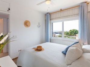 A bedroom with a bed, a window with curtains, a ceiling fan, and a view of the sea. - Apartment  by Interhome (Portocolom)