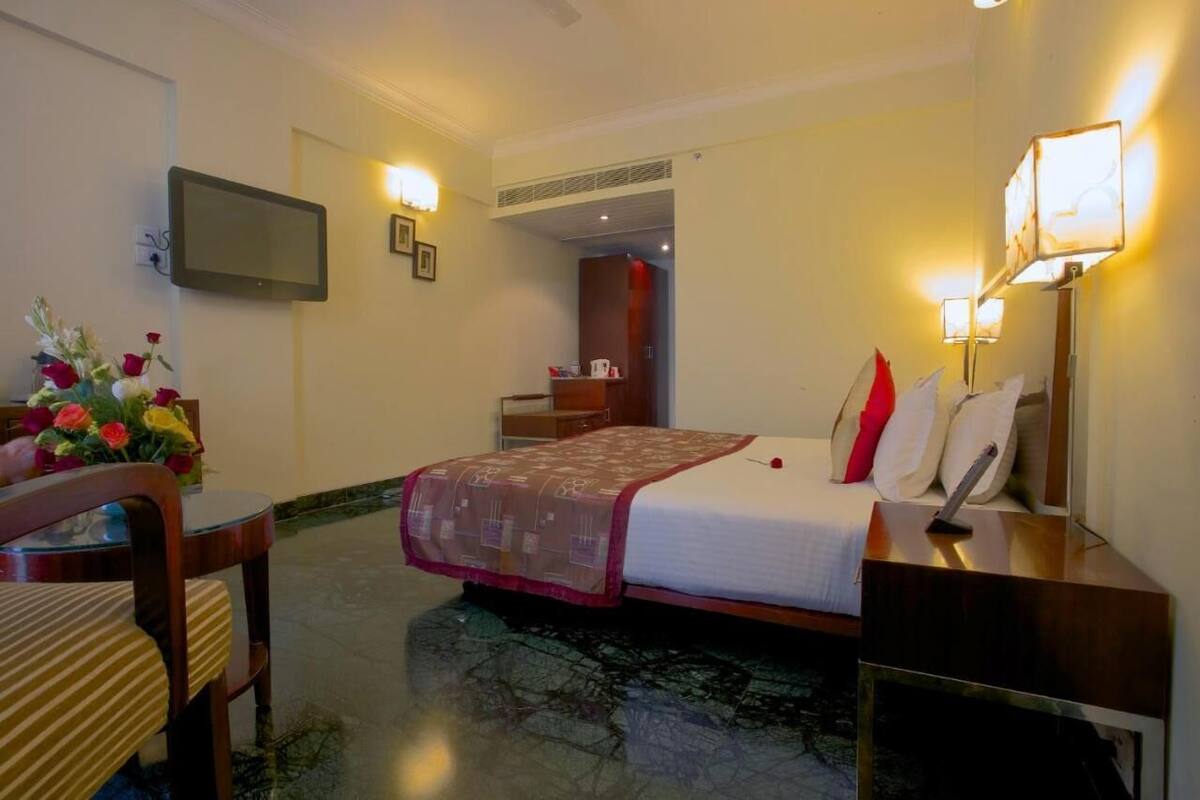 A hotel room with a bed, bedside table, lamp, TV, and a desk with a chair.