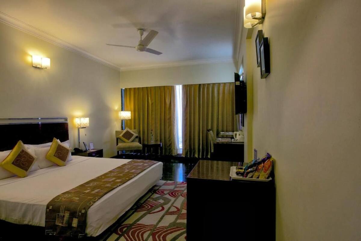 A hotel room with a bed, bedside lamps, a desk, a chair, a ceiling fan, and a window with curtains.