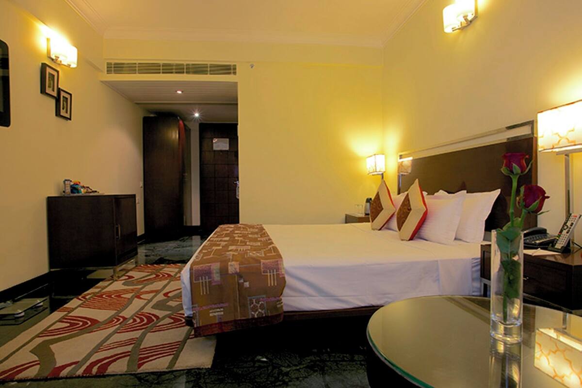 A hotel room with a bed, bedside table, lamp, and a vase with a flower.
