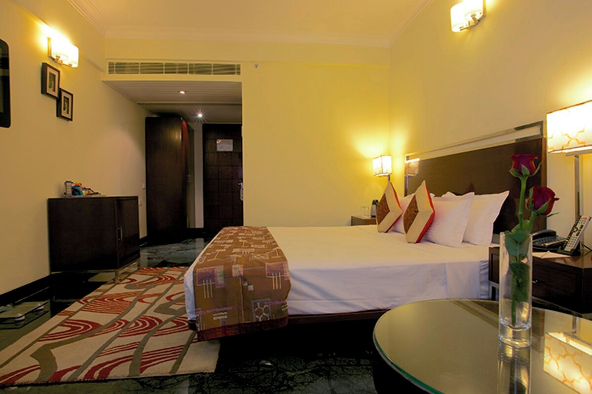 A hotel room with a bed, bedside table, lamp, and a vase with a flower.
