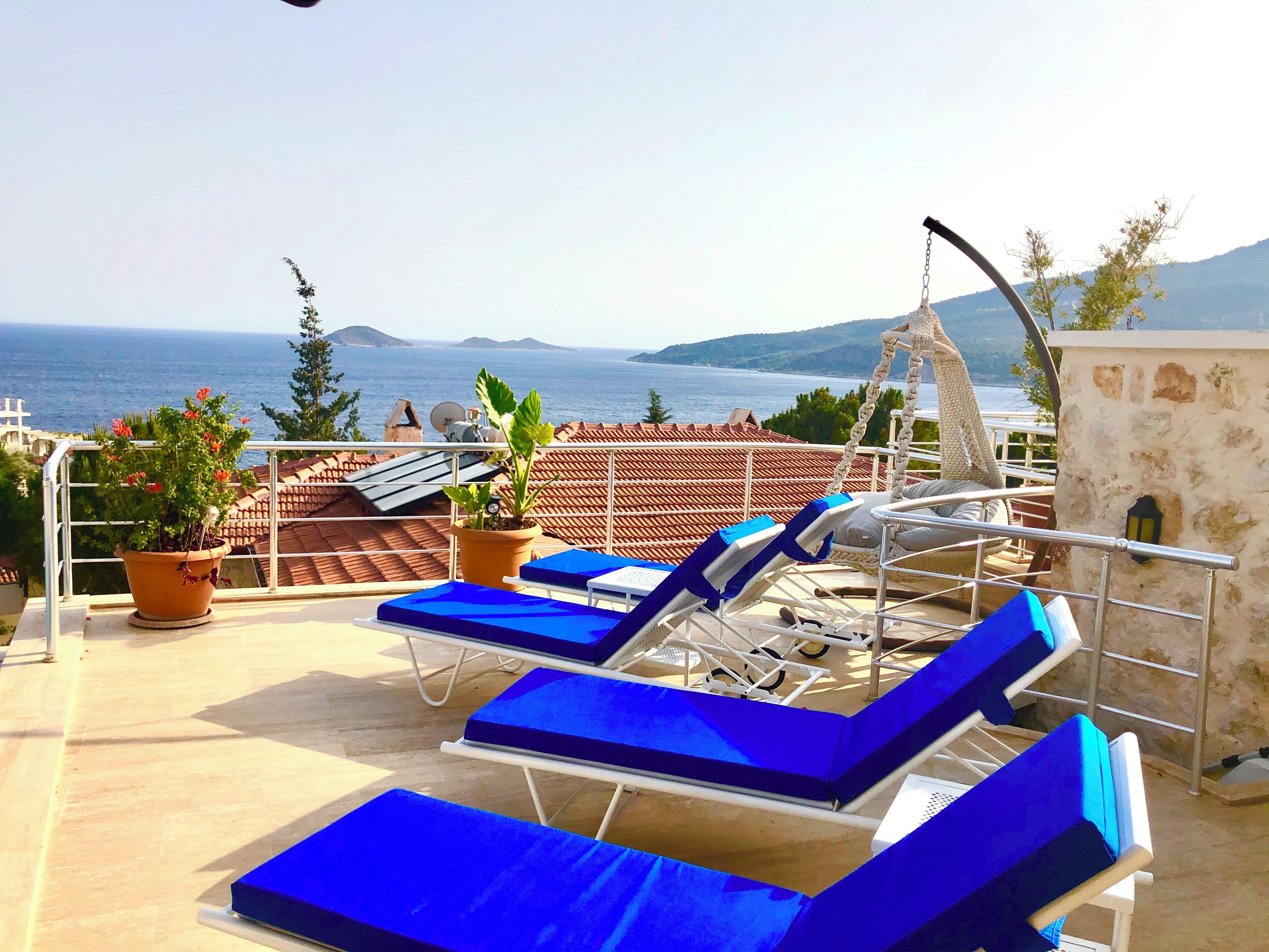 A rooftop terrace with blue lounge chairs, potted plants, and a view of the sea and mountains.