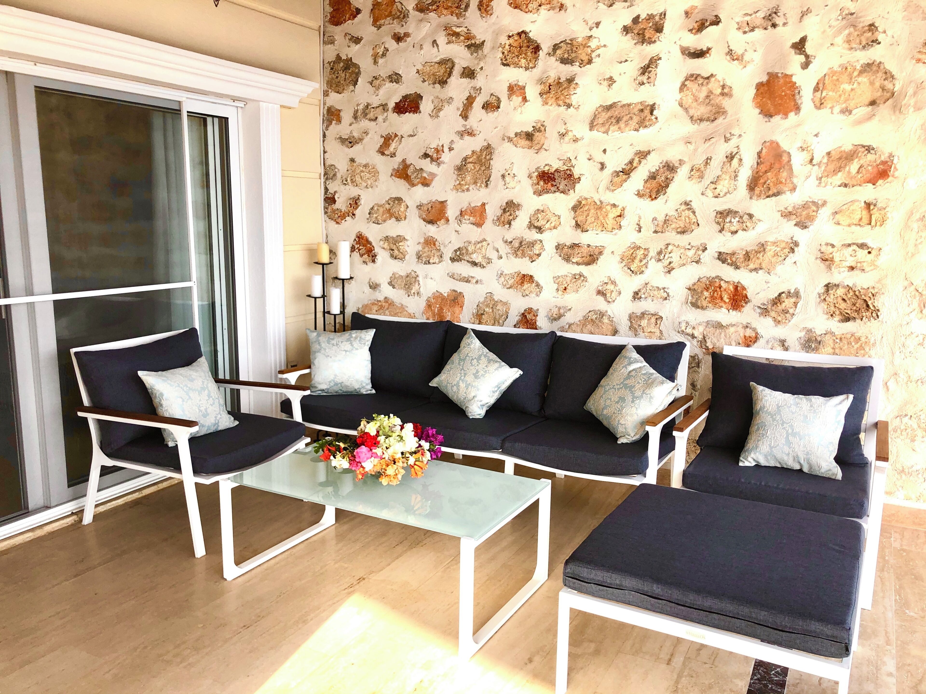 Un salon moderne avec un mur en pierre, un ensemble de canapés, deux fauteuils, une table basse en verre et un arrangement floral en guise de décoration centrale.
