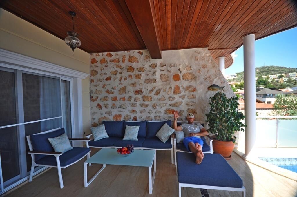 Un espacio de estar al aire libre techado, con pared de piedra, un sofá, dos sillones, una mesa con fruta y una planta en maceta.