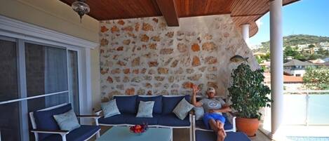 Un espacio de estar al aire libre techado, con pared de piedra, un sofá, dos sillones, una mesa con fruta y una planta en maceta.