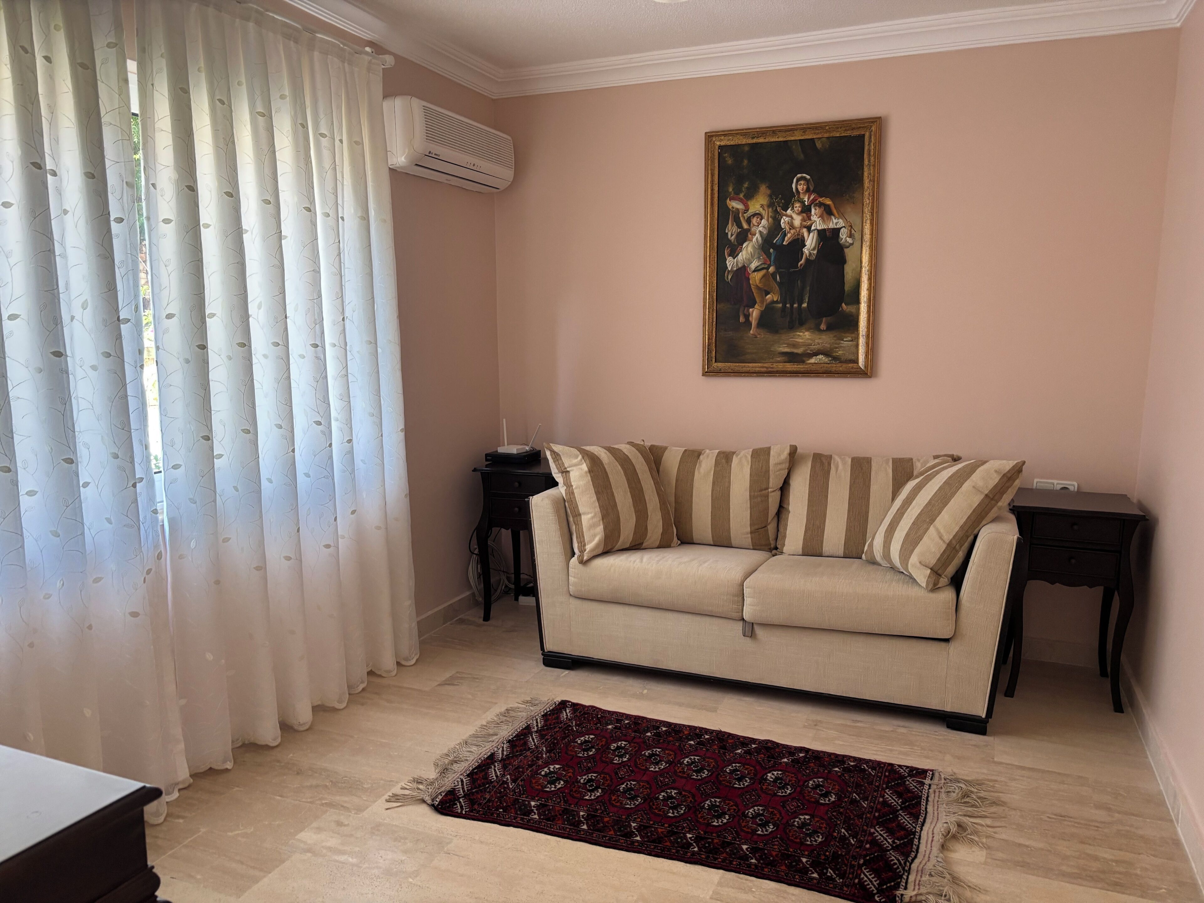 A living room with a beige sofa, a patterned rug, a painting on the wall, and an air conditioning unit.