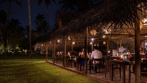 An outdoor dining area with thatched-roof tables and chairs, surrounded by palm trees. - Marari Beach Resort - A CGH Earth Experience (Cherthala)