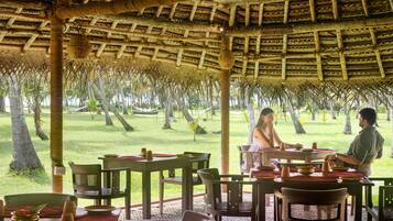Un área de comedor al aire libre con techo de paja, mesas y sillas de madera, rodeada de follaje.