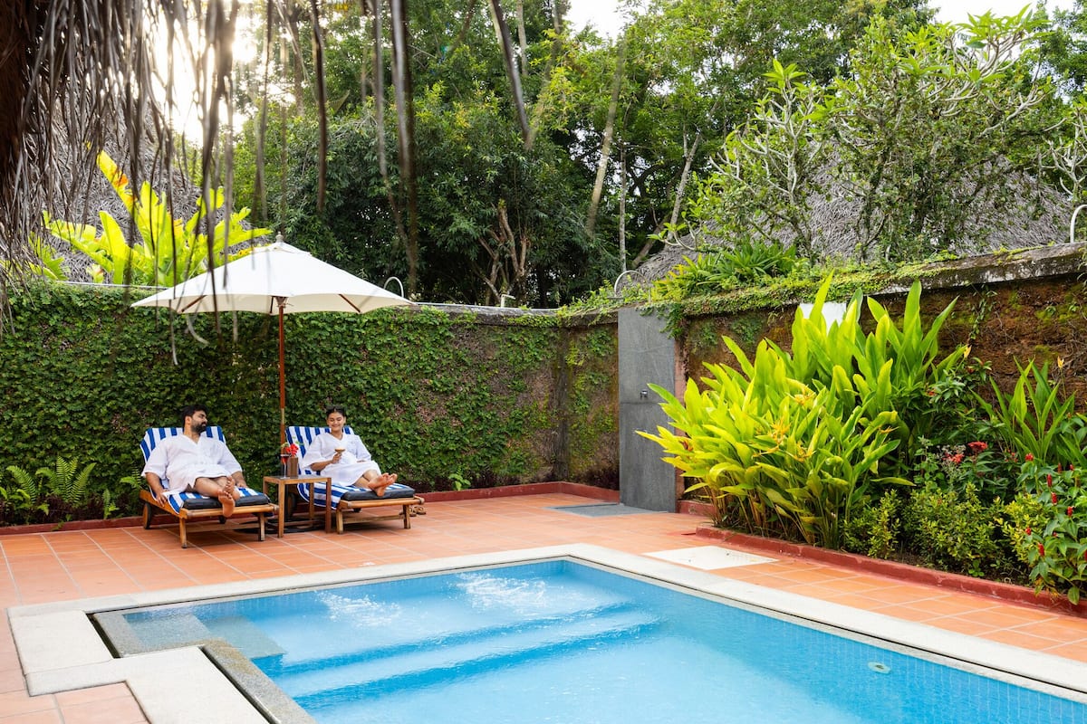 A pool area with two lounge chairs and an umbrella, surrounded by lush greenery and a tiled floor.