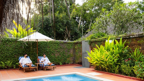 A pool area with two lounge chairs and an umbrella, surrounded by lush greenery and a tiled floor. - Marari Beach Resort - A CGH Earth Experience (Cherthala)