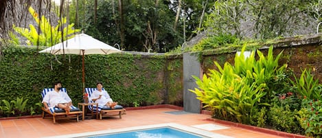 A pool area with two lounge chairs and an umbrella, surrounded by lush greenery and a tiled floor.