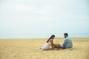 A beach with two people sitting on a blanket, a basket, and a bottle.