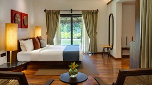 A hotel bedroom with a bed, bedside lamps, a round table with a vase, a view of greenery through the window, and a ceiling fan. - Marari Beach Resort - A CGH Earth Experience (Cherthala)