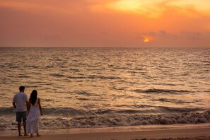 A couple standing on a beach facing the ocean at sunset.